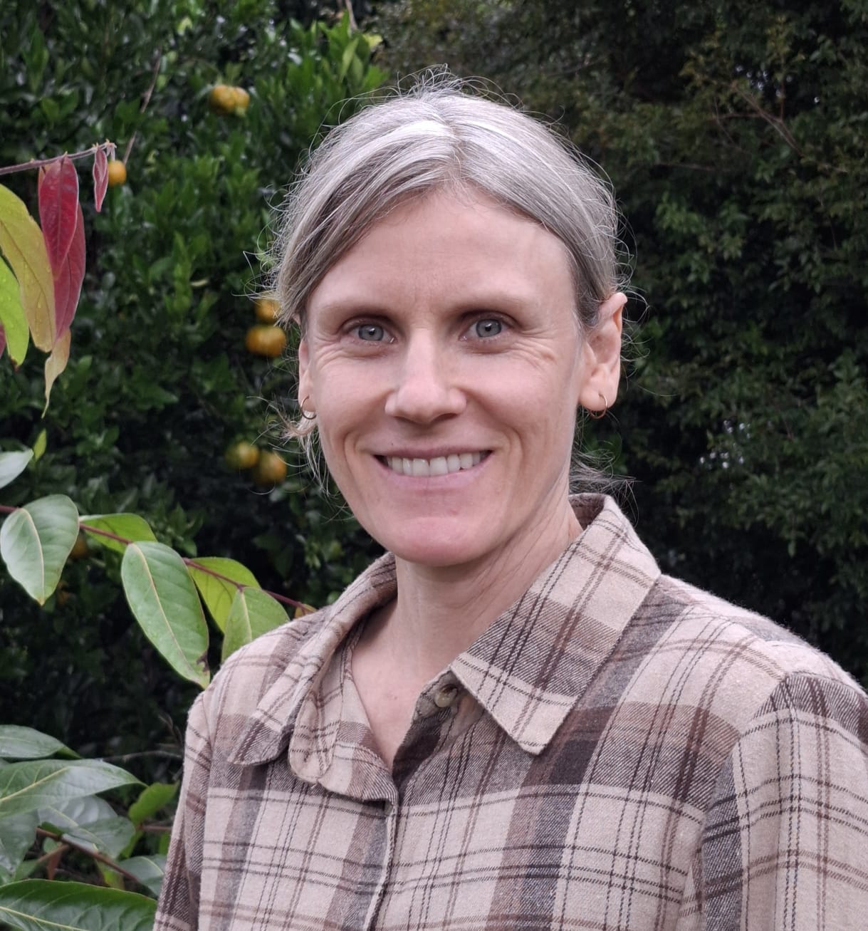 Woman with grey hair smiling outdoors, wearing plaid shirt with leafy garden background.